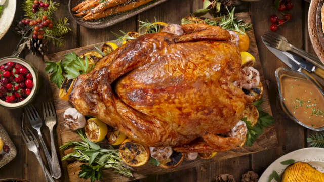 A holiday feast: a roasted turkey on a wooden board, surrounded by herbs, garlic, mushrooms, veggies, and gravy.