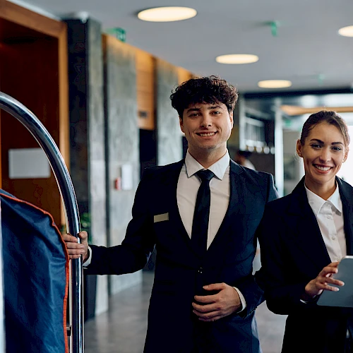 Two hotel staff in uniform stand near a luggage cart, smiling; a man in suit and a woman with a tablet greet guests at the lobby.