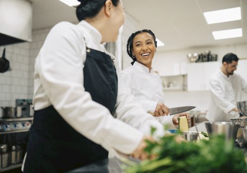 Two chefs in a busy kitchen share a smile while prepping ingredients and chatting near a workstation with bowls and herbs.