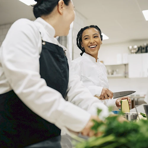 Two chefs in a busy kitchen share a smile while prepping ingredients and chatting near a workstation with bowls and herbs.