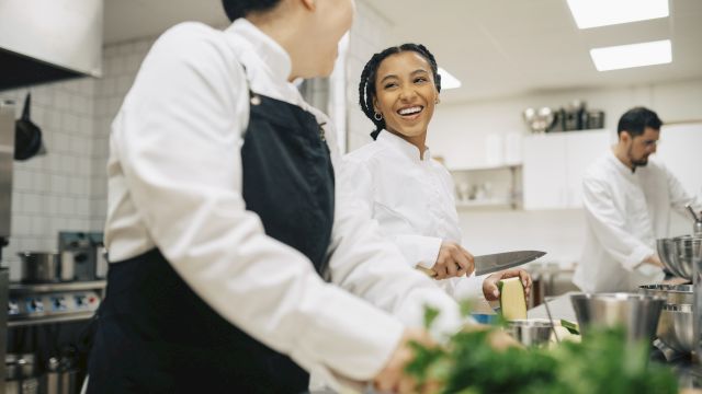 Two chefs in a busy kitchen share a smile while prepping ingredients and chatting near a workstation with bowls and herbs.