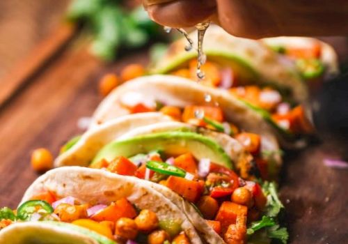 A hand squeezes lime over a row of tacos filled with chickpeas, avocado, and vegetables on a wooden board, garnished with lime slices.