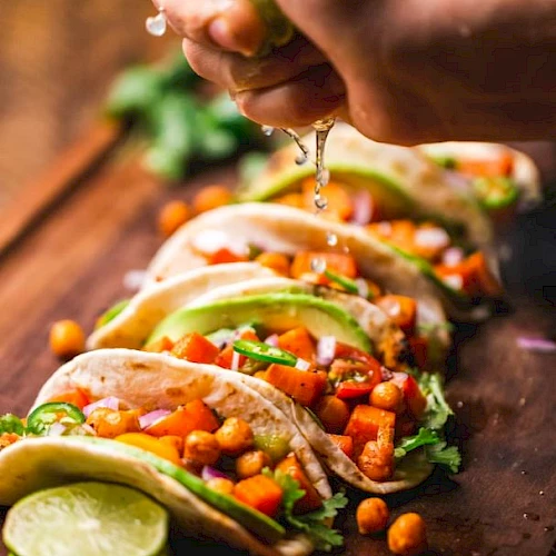 A hand squeezes lime over a row of tacos filled with chickpeas, avocado, and vegetables on a wooden board, garnished with lime slices.