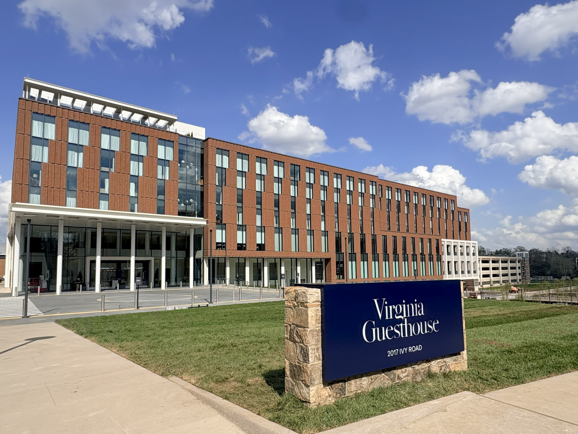 A modern brick dormitory building with large windows and a blue &ldquo;Virginia Guesthouse&rdquo; sign at the entrance, under a bright, partly cloudy sky.