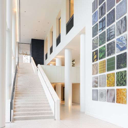 A bright, modern interior with a wide staircase, tall white walls, and a colorful mosaic wall display of small square tiles on the right.