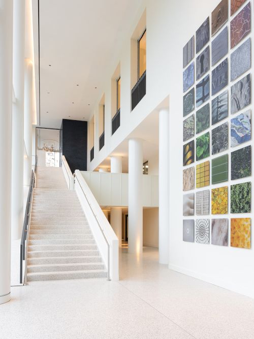 A bright, modern interior with a wide staircase, tall white walls, and a colorful mosaic wall display of small square tiles on the right.