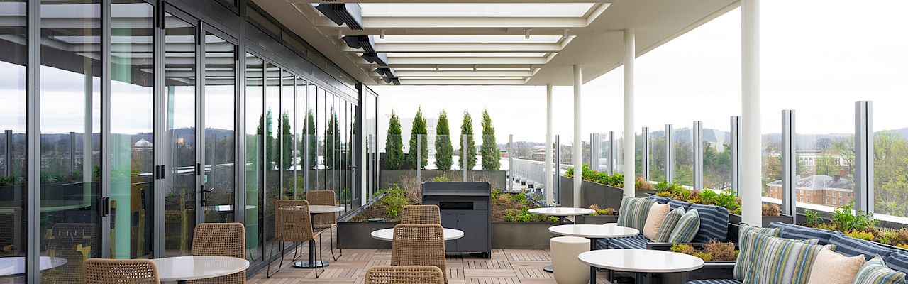 A modern outdoor rooftop seating area with wicker chairs, round tables, and cushioned couches under a covered glass railing terrace, sunny view ahead.