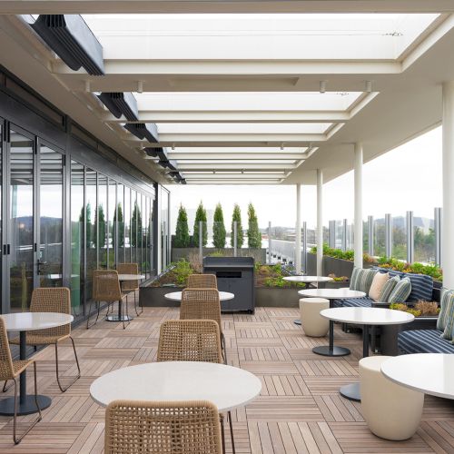 A modern outdoor rooftop seating area with wicker chairs, round tables, and cushioned couches under a covered glass railing terrace, sunny view ahead.