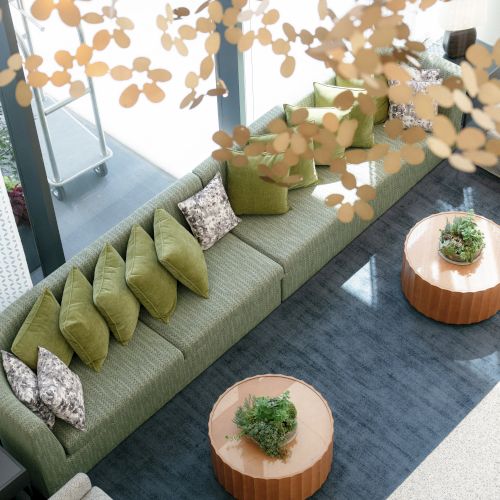 A cozy green sofa lounge with pillows, wooden side tables with small plants, and hanging gold flower cutouts in a bright indoor setting.