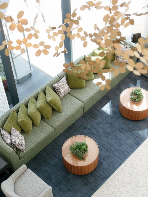 A cozy green sofa lounge with pillows, wooden side tables with small plants, and hanging gold flower cutouts in a bright indoor setting.