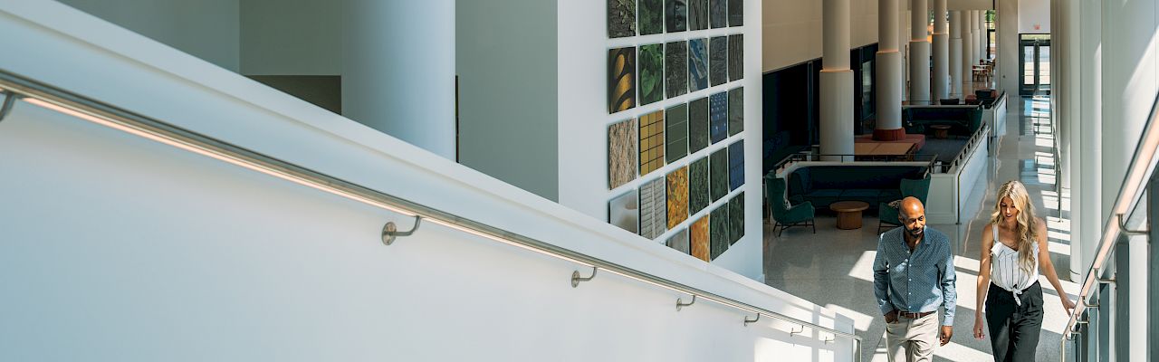 A bright modern building interior shows a staircase, colorful art wall panels, and two people walking toward a lobby.