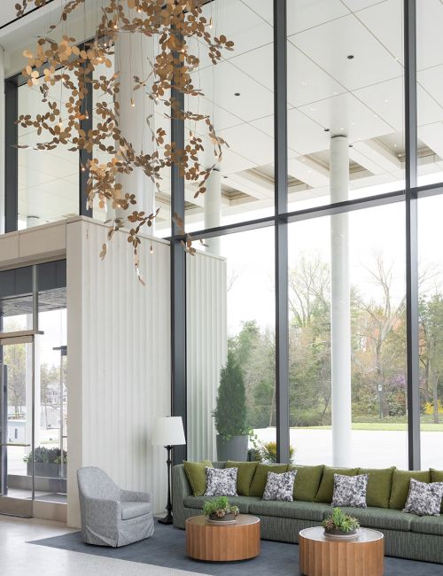 A modern hotel lobby with a high glass wall, green sectional sofa, round wooden coffee tables, plants, and a hanging branch chandelier.