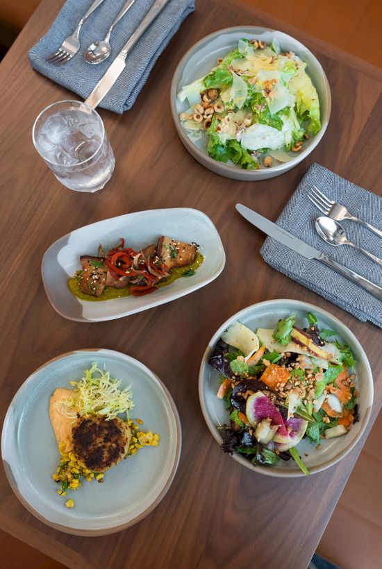 A colorful spread of bowls and a small plate with salads, toppings, and a grilled veggie skewer on a wooden table, ready to share.