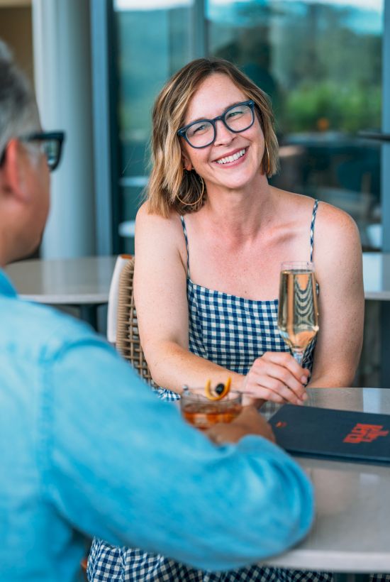 A smiling woman in glasses at an outdoor patio table clinks drinks with someone, with a relaxed sunny backdrop.