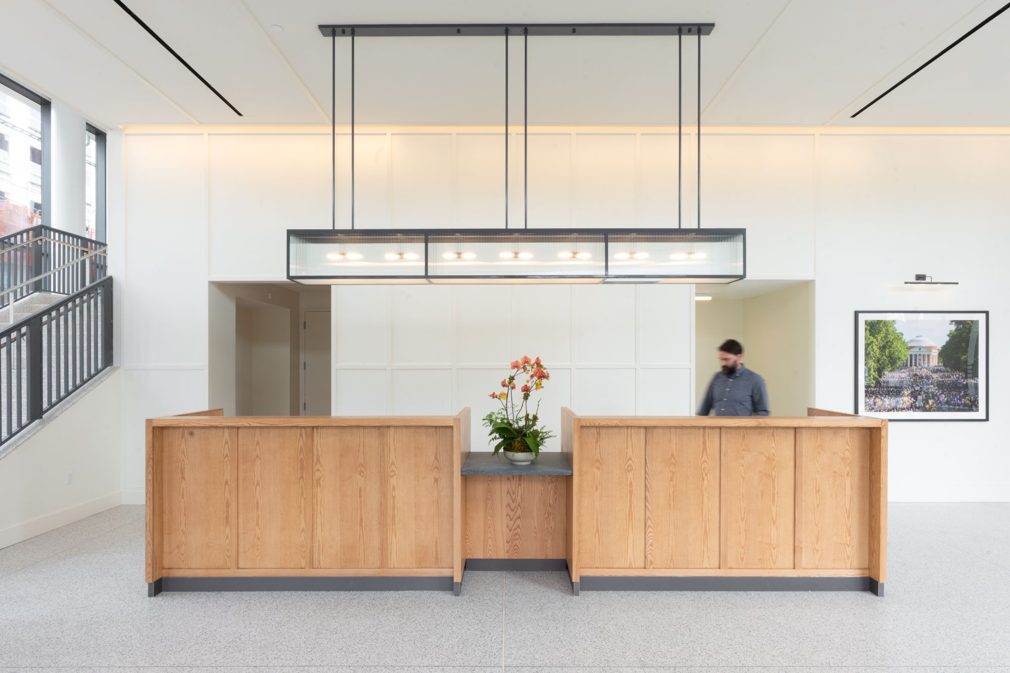 A bright modern lobby shows a wooden reception desk, hanging light fixture, wall art, and a man walking in the background.