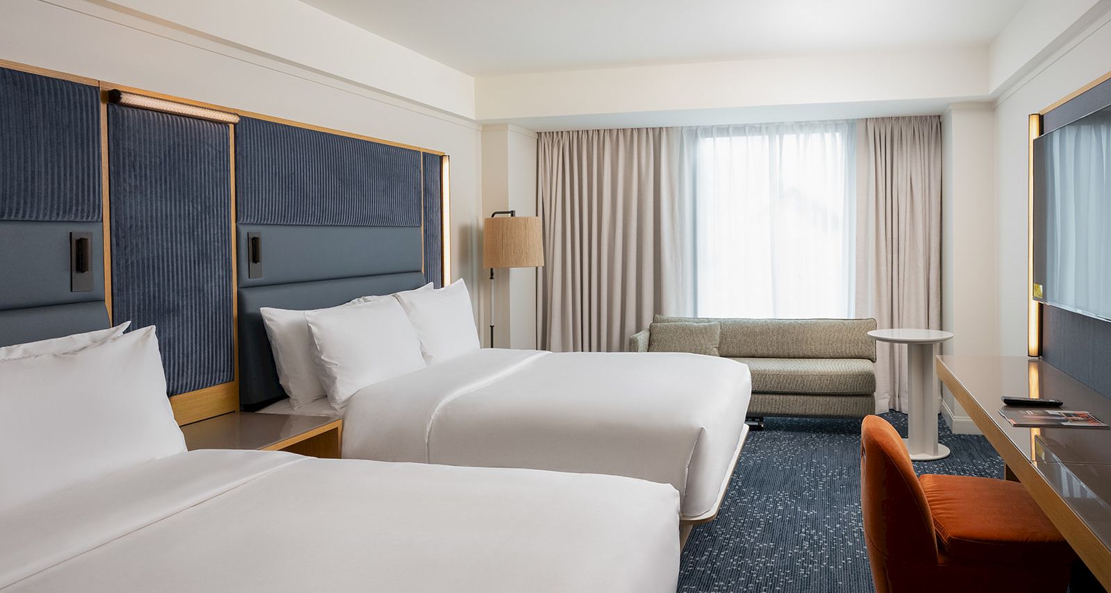 A modern hotel room with two large white beds, blue padded headboards, a sofa by a window, a desk with an orange chair, and soft beige curtains.