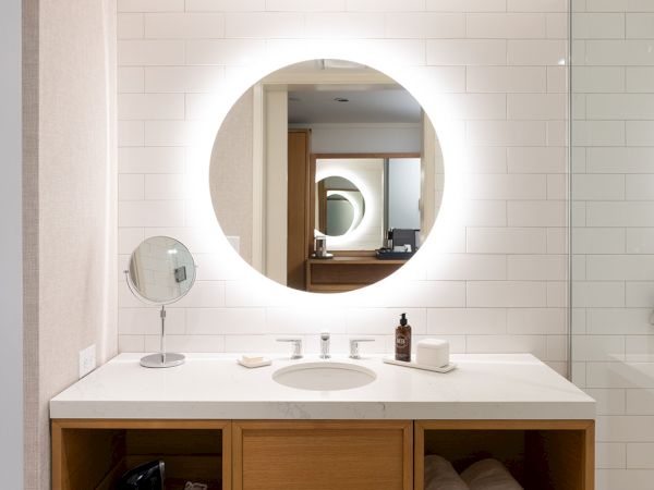 A modern bathroom vanity with a round backlit mirror, white countertop, wooden cabinet drawers, towels, and sink accessories.