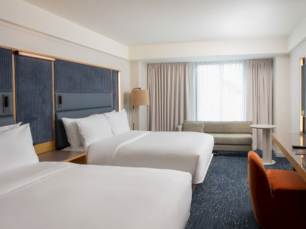 Two hotel beds with white linens, a blue padded headboard wall, a sofa by a window, and a desk with an orange chair in a bright room.