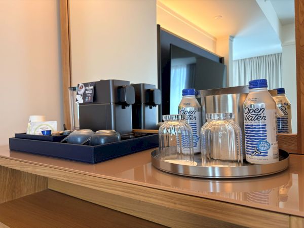 A hotel coffee/water station with dispensers and several &ldquo;open water&rdquo; bottles on a tray, all in a bright room.