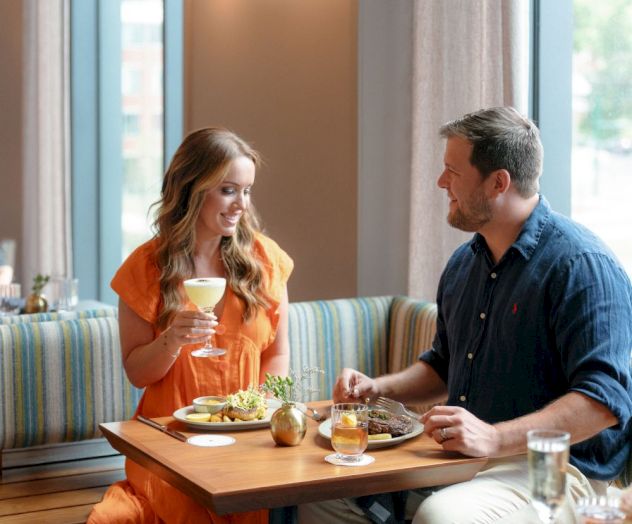 A woman in orange and a man in blue dine together at a table, smiling over drinks and plates in a cozy restaurant.