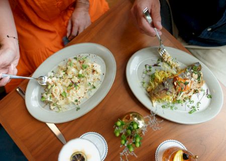 Two plates of gourmet food on a wooden table as someone serves a dish with a spoon and forks.