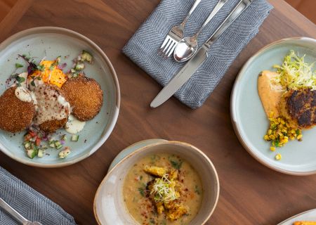A top-down view of three plates with assorted plated dishes: croquettes with salad, a creamy soup, and a crispy item with greens, on a wooden table.