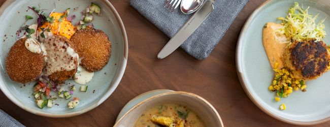 Two plated entr&eacute;es and a bowl of soup sit on a wooden table with forks and napkins.