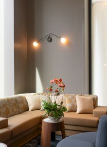 A cozy living room with a tan sectional, a round wooden coffee table, a plant with orange flowers, soft cushions, and warm wall lights.