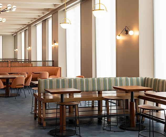 A stylish dining area with long booths, round wooden tables, and pendant lights along tall windows, creating a bright, airy space.