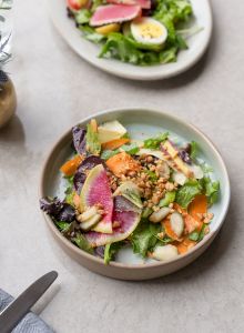 A bowl of mixed green salad topped with sliced radishes, carrots, cucumber, beets, avocado, and crunchy grains.