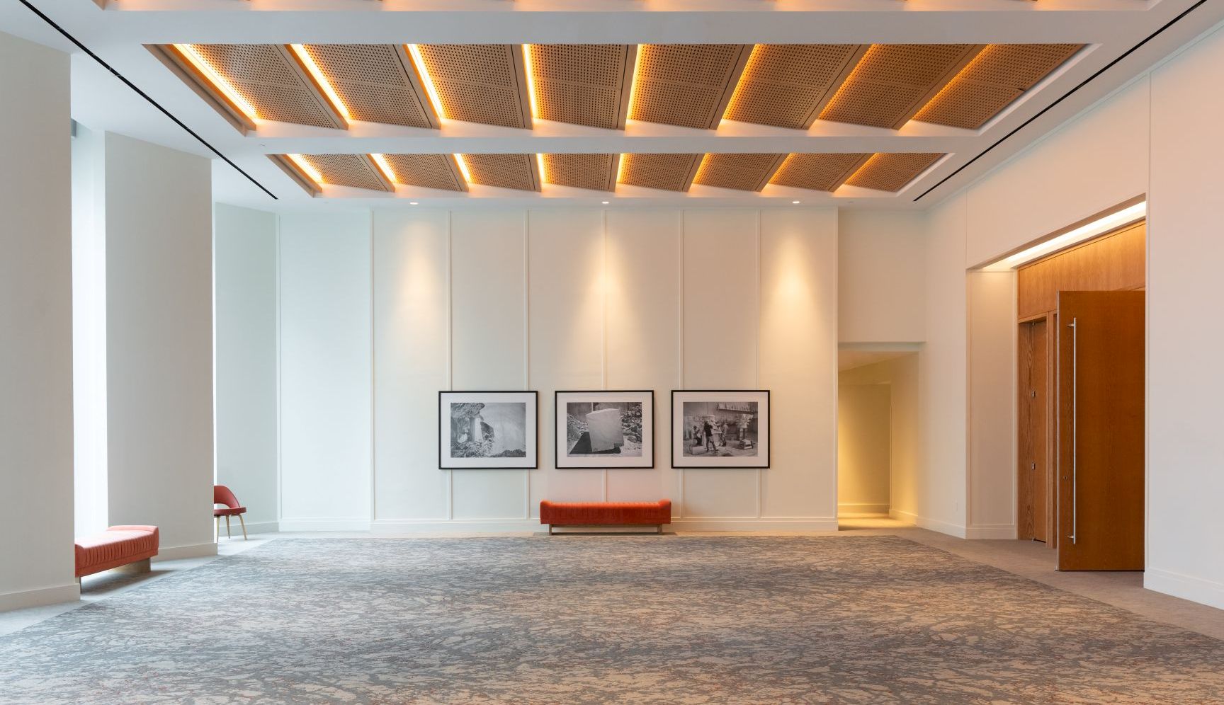 A modern gallery room with recessed warm lights, white panels, framed photos, an orange bench, and polished carpet, in view.