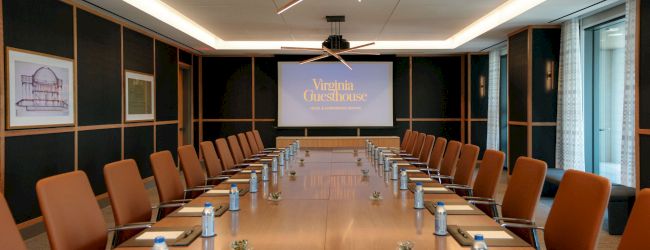 A conference room with a long table set with water bottles, chairs, and a projection screen reading &ldquo;Virginia Guesthouse&rdquo;.