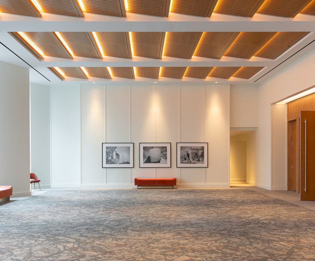 A spacious, modern gallery with white walls, framed artwork, wooden ceiling accents, and a gray patterned carpet, plus red benches and soft lighting.