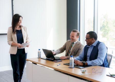 A woman stands in a bright office, holding papers while three coworkers sit and smile around laptops during a meeting.