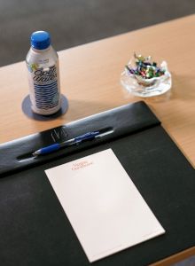 A desk setup with a bottled water drink, blue-and-black pen, a small bowl of colorful candies, and a &ldquo;Virginia Ghicehouse&rdquo; notepad.