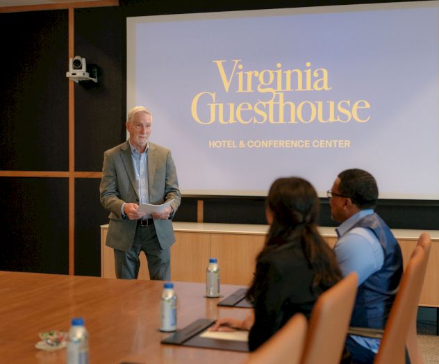 A man presents to an audience in a conference room, with a screen reading &ldquo;Virginia Guesthouse Hotel & Conference Center&rdquo; behind him.
