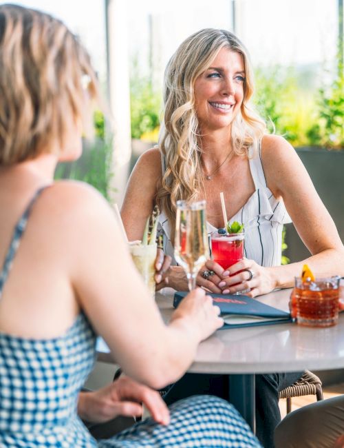 Two women chat at an outdoor patio, smiling and toasting with drinks, one holding a red cocktail with a straw.