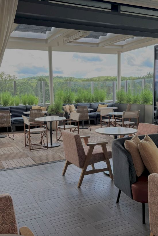 A modern covered patio with cushioned seating, round tables, and large windows overlooking lush greenery in daylight.