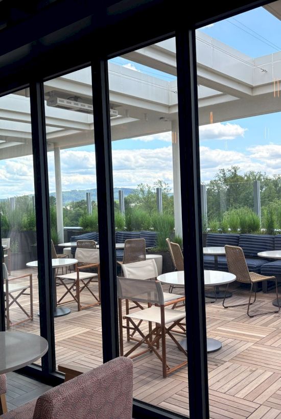 View of a modern patio with round tables and woven chairs through large glass doors, overlooking greenery and a cloudy sky.