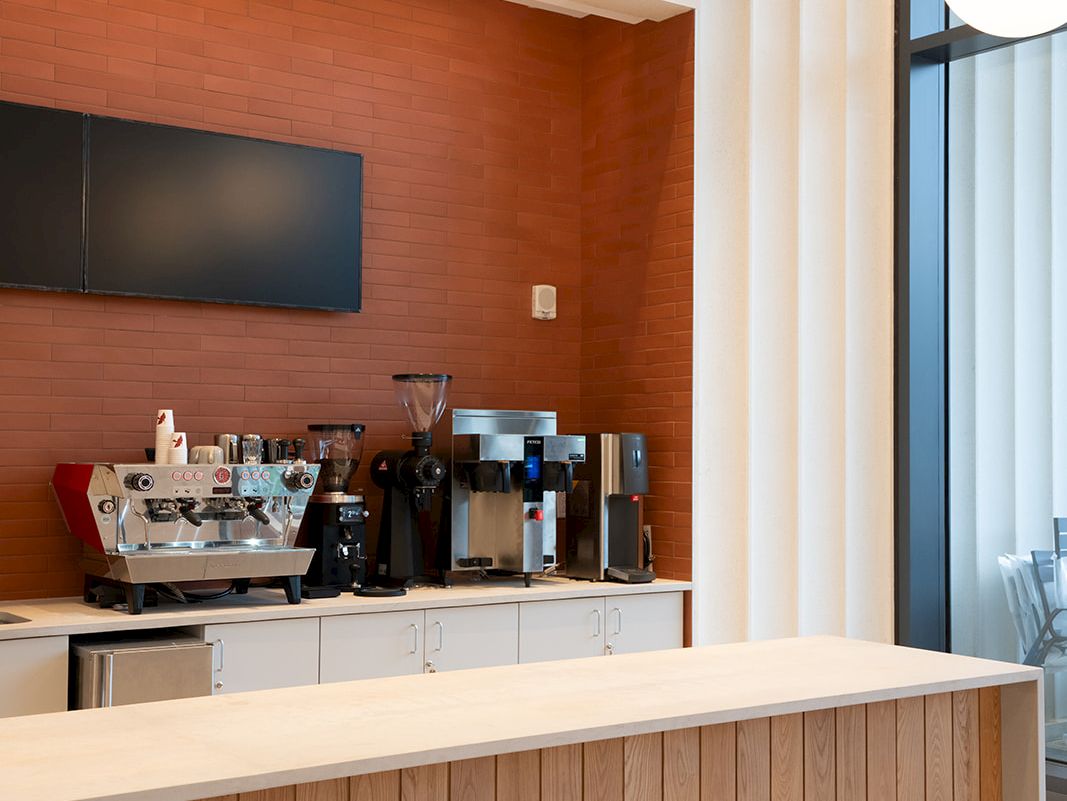 A modern cafe counter with a wooden front, coffee machines, a wall-mounted TV, and large windows with vertical panels, topped by round pendant lights.