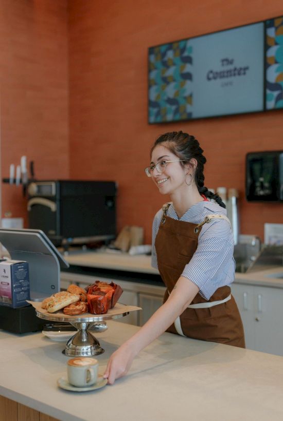 Smiling barista in a cafe behind the counter, serving coffee with pastries on a stand.