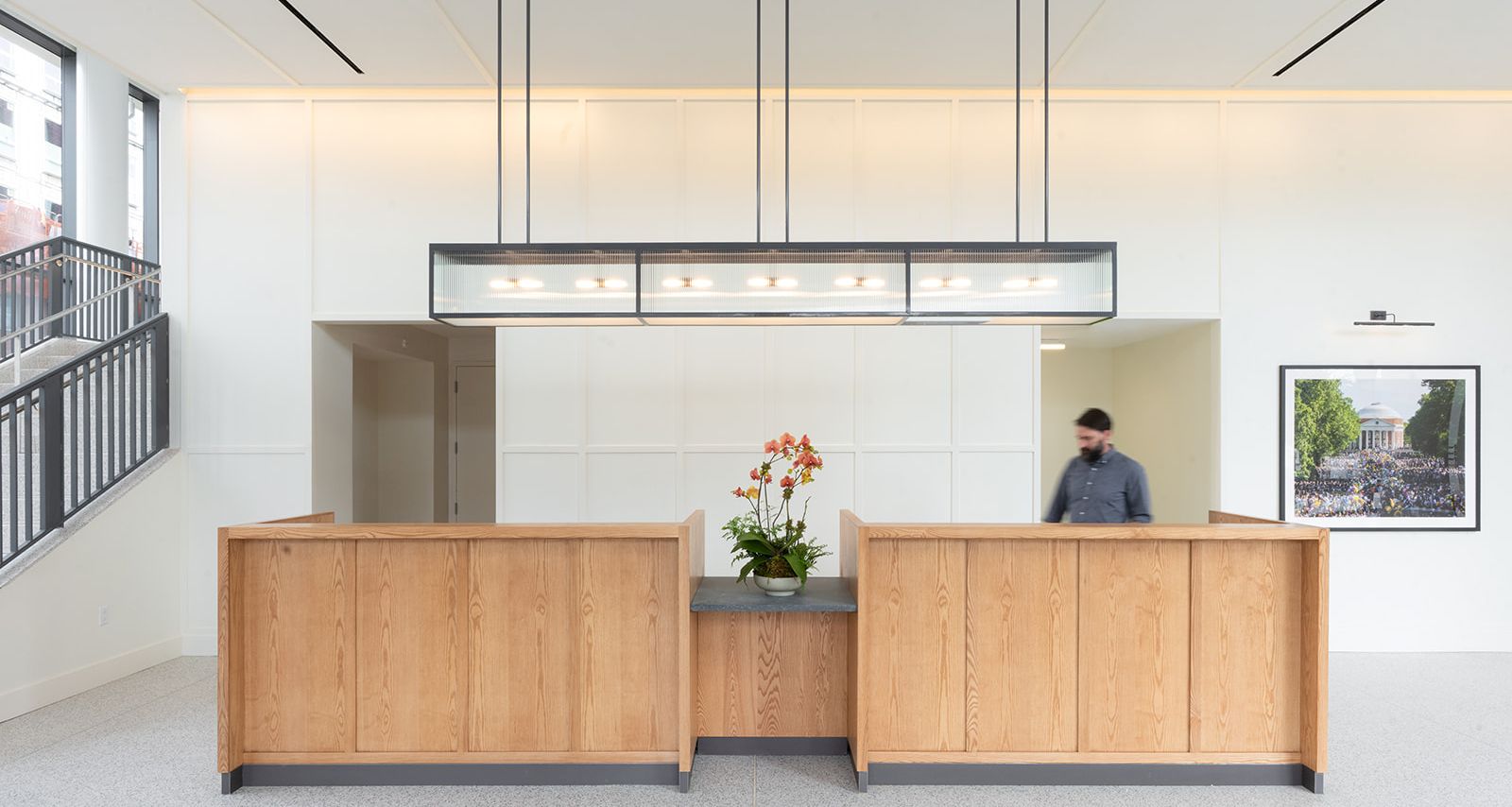 A modern office reception area with two wooden front desks, a central flower arrangement, a suspended rectangular light fixture, and a person standing near a wall with framed artwork.