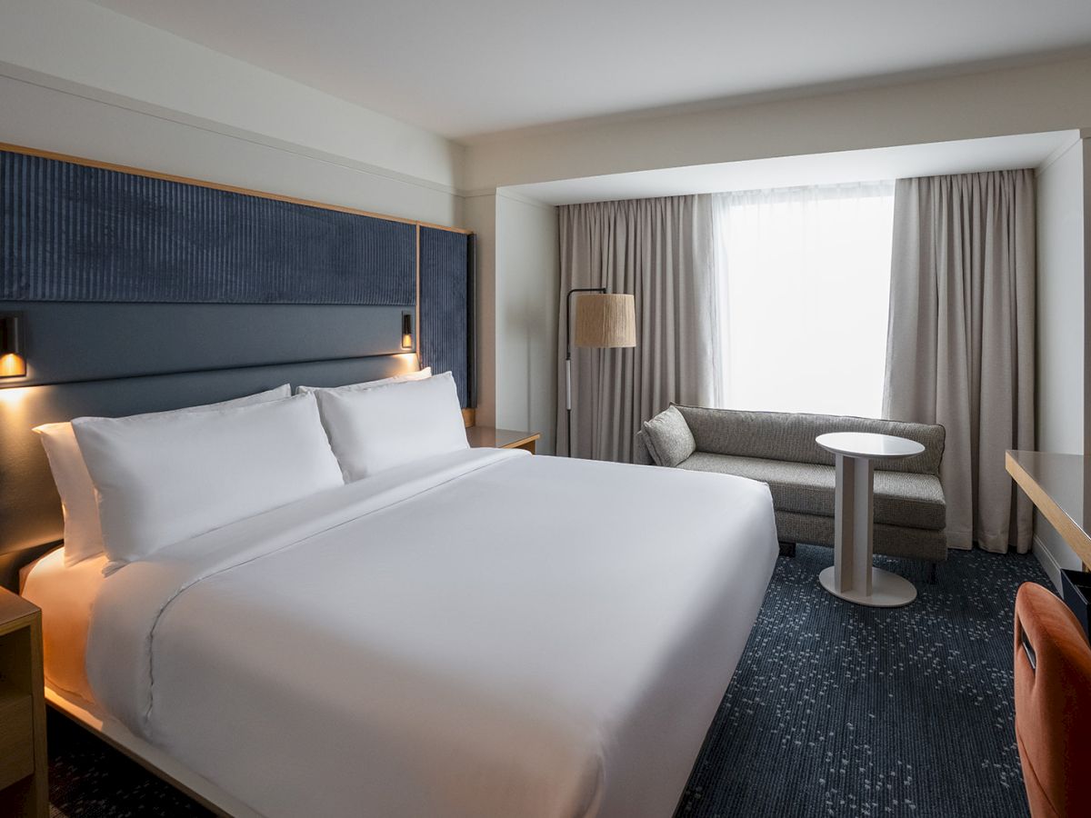 A modern hotel room with a large bed, navy headboard, two bedside lamps, a seating area by a window, and neutral tones&mdash;clean and cozy.