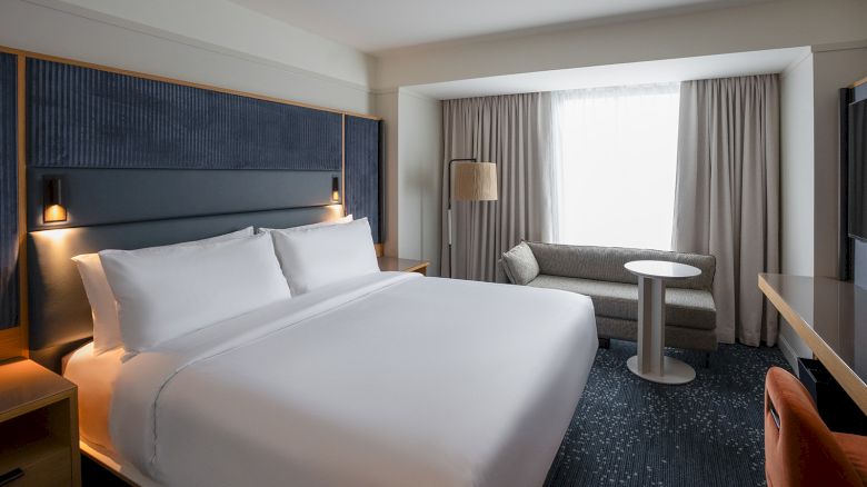A modern hotel room with a large bed, navy headboard, two bedside lamps, a seating area by a window, and neutral tones&mdash;clean and cozy.