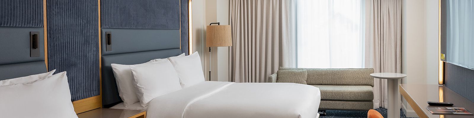 Two neatly made twin beds in a modern hotel room, with a cushioned headboard, a sofa by a window, a desk with an orange chair, and soft lighting, ending with a period.