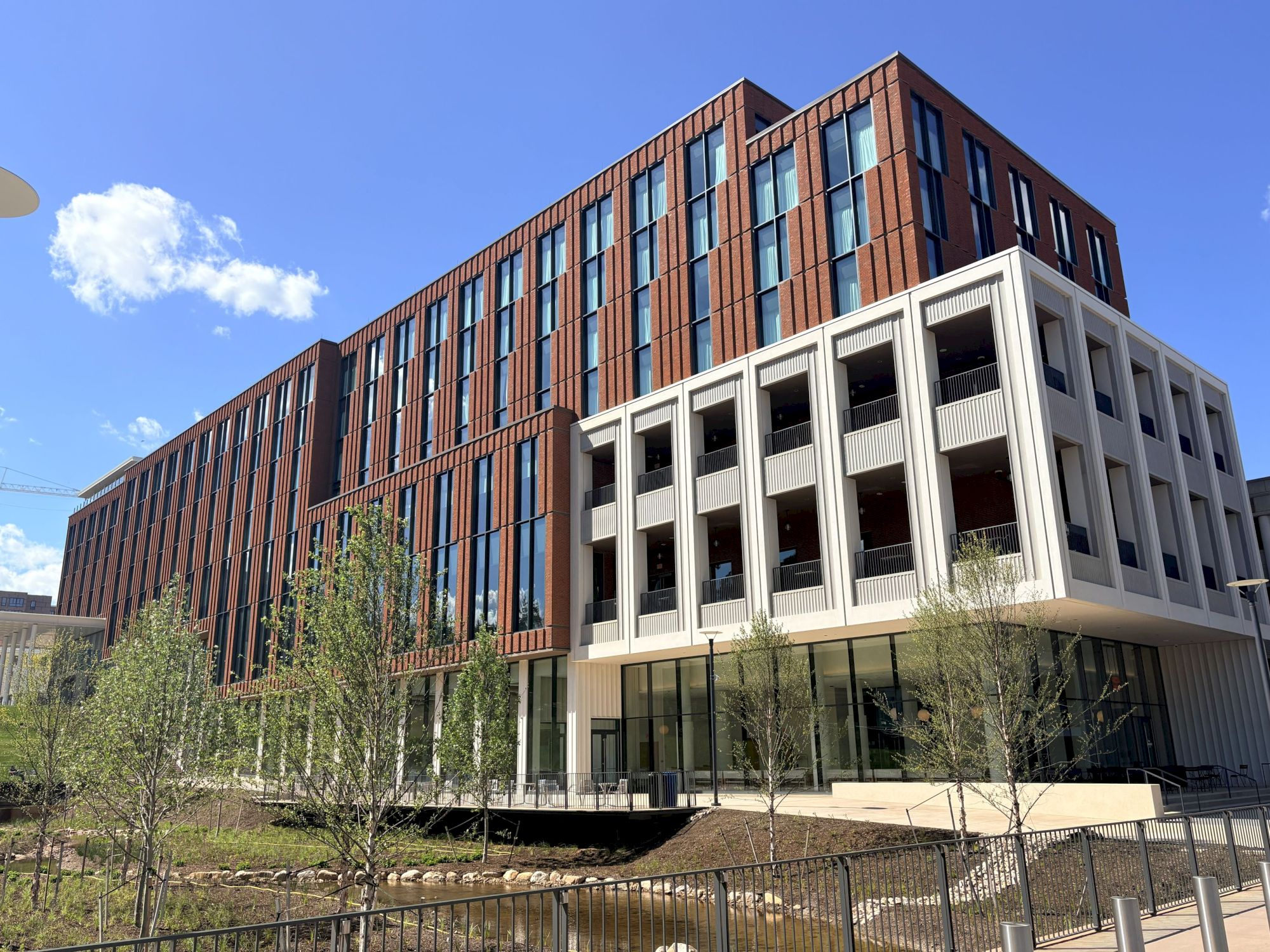 A modern brick-and-glass apartment building with balconies stands against a clear blue sky, with trees in the foreground.