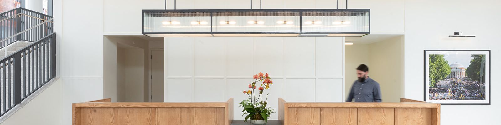 A modern office lobby with a wooden reception desk, hanging lights, flowers, and a framed photo on the wall.