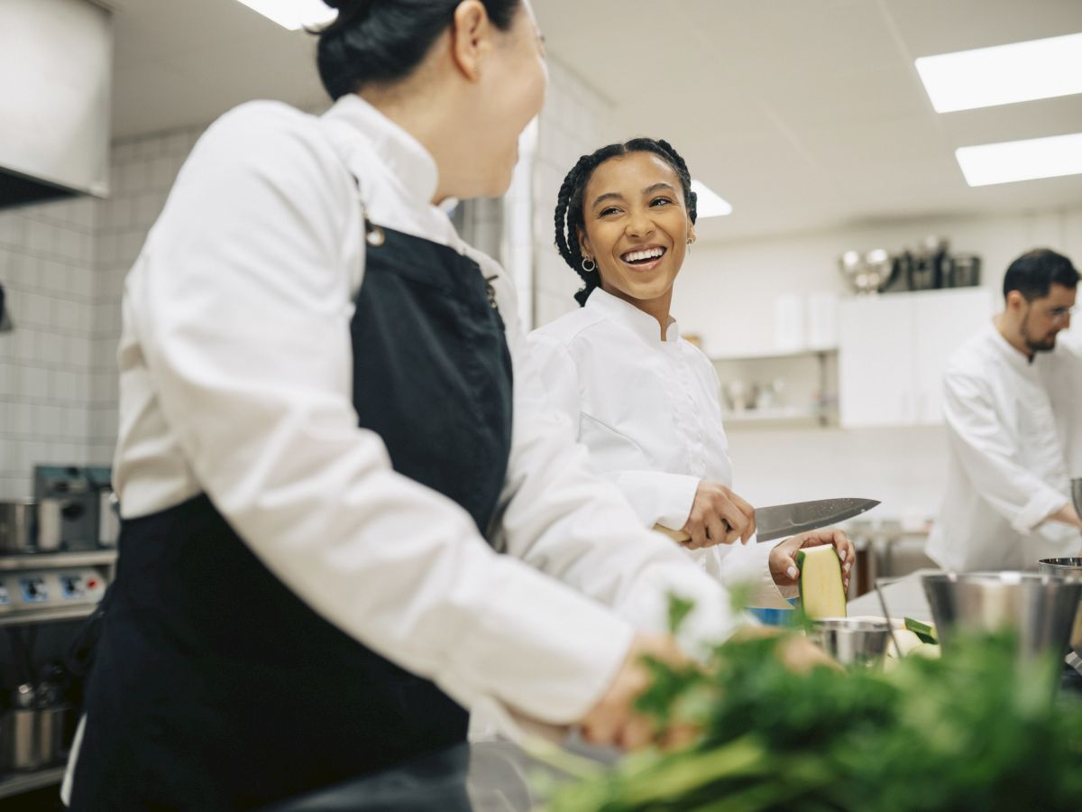 In a bright kitchen, chefs in white jackets work together as one smiles while slicing ingredients with a knife.