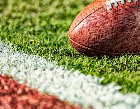 A close-up of a brown football on a grassy field near a white sideline, with a blurred red track in the foreground.
