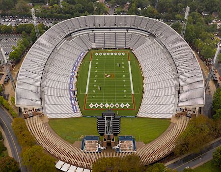 An aerial view of a large football stadium with a green field, white yard lines, and surrounding seating filled with light-colored seats, all amid trees and roads.